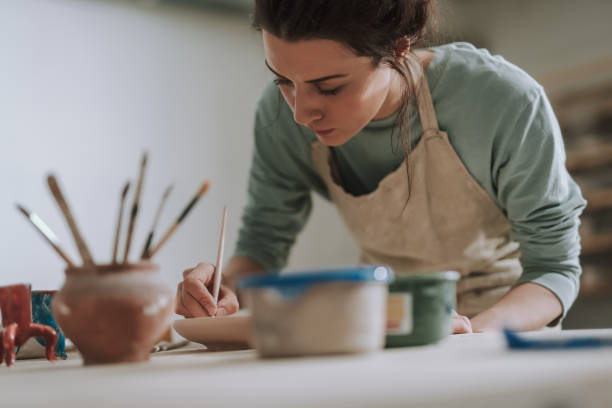 Skillful young woman in apron painting pottery at workshop Close up portrait of beautiful brunette lady drawing on clay plate while leaning over the table with pottery and paints artist stock pictures, royalty-free photos & images