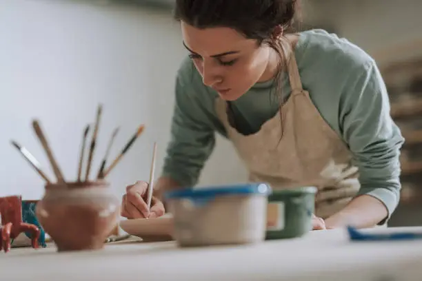 Close up portrait of beautiful brunette lady drawing on clay plate while leaning over the table with pottery and paints