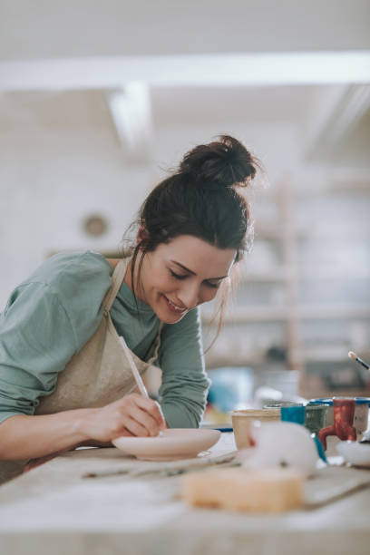 Cheerful young woman painting pottery at workshop Beautiful brunette lady in apron drawing on ceramic plate and smiling pottery photos stock pictures, royalty-free photos & images