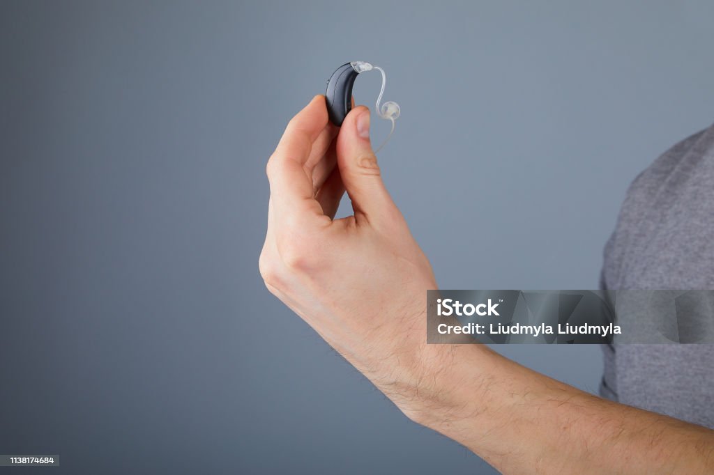 holding small black hearing aid Man holding small black hearing aid, closeup. Hearing aids on grey background, alternative to surgery. Hearing Aid Stock Photo