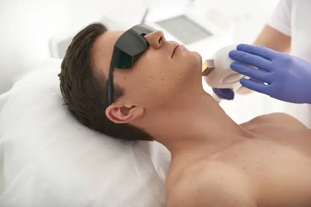 Beauty esthetics treatment. Close up side on portrait of young attractive male having laser hair removal procedure of chin zone in cosmetic center