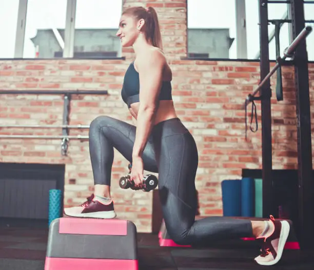 Photo of Cheerful athlete woman doing lunges with dumbbells on a support in the gym. Training concept