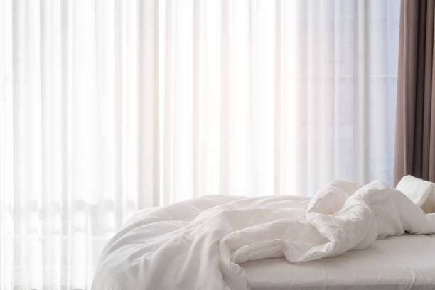 Unmade bed with shades of sunlight through the curtain in morning. stock photo