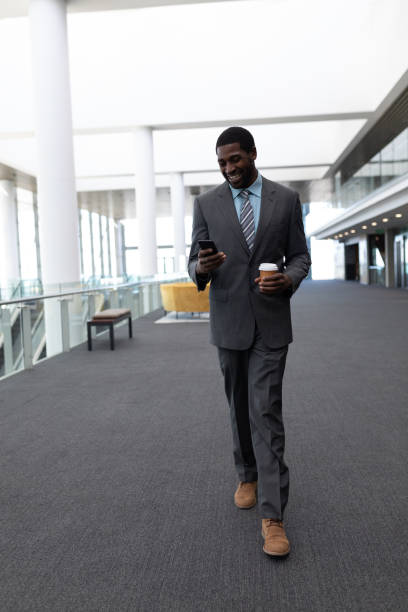 istock Afrrican-american businessman with coffee cup using mobile phone while walking in office 1138206314
