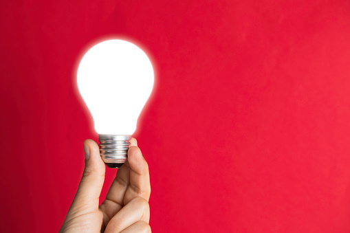Hand is holding bright light bulb in front of red background.