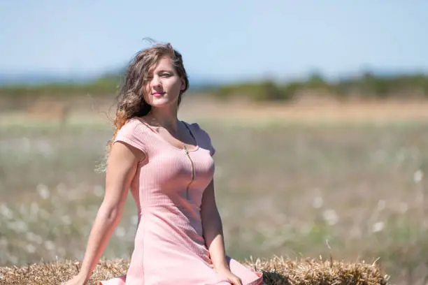 Countryside landscape in Tuscany, Italy with young girl woman sitting on hay bale in dress with hair in wind posing model