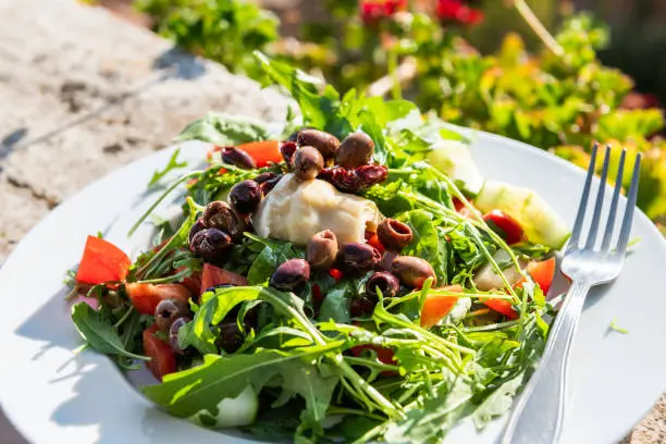 Closeup of arugula salad with olives and artichoke on plate on balcony terrace by red geranium flowers in garden outside in Italy in Tuscany