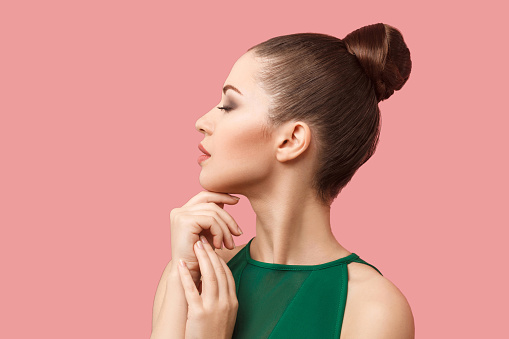 Profile side view portrait of calm serious beautiful young woman with bun hairstyle and makeup in green dress standing with closed eyes and touching her chin.