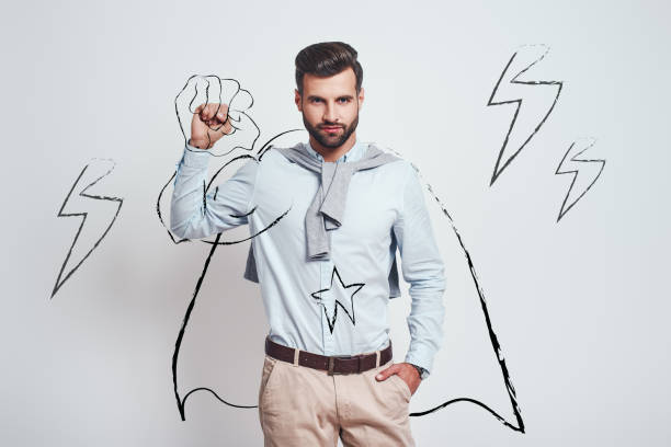 You can do it! Young attractive brunet wearing a drawn cape and raising one hand clanched in fist above his head while standing against grey background with illustration of the lightning bolts You can do it! Young attractive brunet wearing a drawn cape and raising one hand clanched in fist above his head while standing against grey background with illustration of the lightning bolts. Success concept superhero photos stock pictures, royalty-free photos & images
