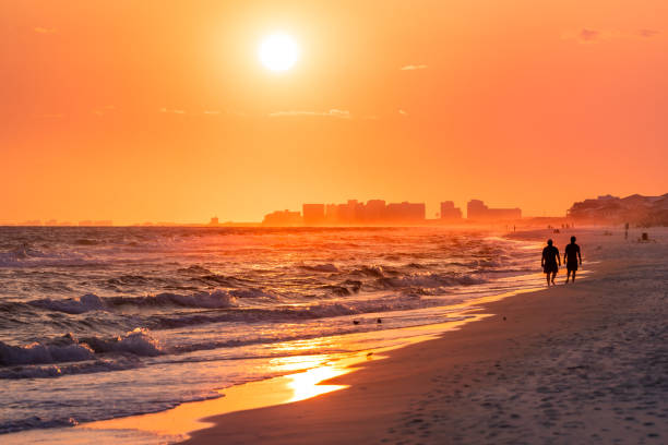 couples marchant sur le rivage avec le coucher du soleil jaune rouge spectaculaire d'orange dans la plage de santa rosa, floride avec la ligne d'horizon de la côte côtière de pensacola en panhandle avec les vagues de océan de golfe du mexique - pensacola photos et images de collection