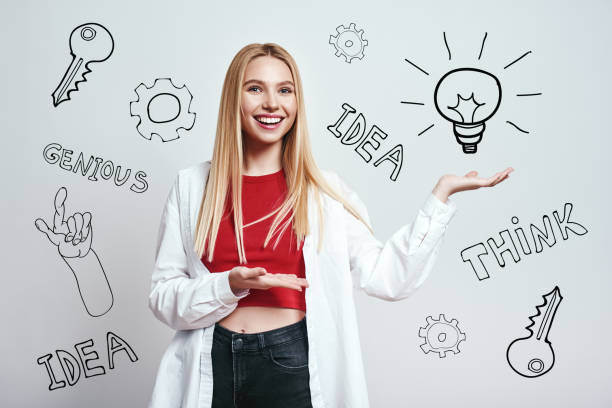 istock Genius idea! Cheerful and young blonde woman showing lightbulb while standing against grey background with hand drawn doodles on it 1138220109