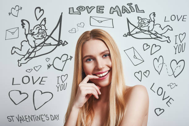 istock Flirty mood. Young female with naked shoulders touching her lips and looking at camera while standing against grey background with doodles on the theme of St.Valentine's day. 1138220315