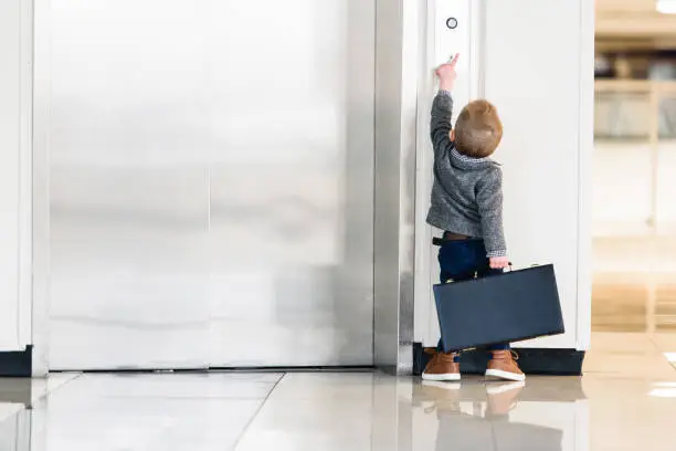 Little boy (preschool aged) in businessman attire, carrying a briefcase, on tippy toes to try and reach the elevator button.