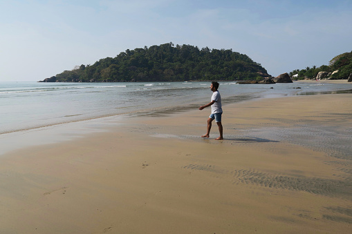 Indian man wearing denim shorts and a blue and white striped t-shirt walking towards sea on the damp compacted sand at the water's edge of Palolem Beach, Goa, India.
