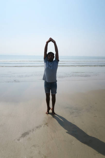 Image of Hindu Indian man practicing Mountain Pose On Tiptoes Arms Flow (Tadasana On Tiptoes Hasta Vinyasa) yoga position on wet sandy beach, water's edge, Palolem Beach, Goa, India Indian man wearing denim shorts and a blue and white striped t-shirt practicing Mountain Pose On Tiptoes Arms Flow (Tadasana On Tiptoes Hasta Vinyasa) yoga position on damp compacted sand at the water's edge of Palolem Beach, Goa, India. palolem beach stock pictures, royalty-free photos & images