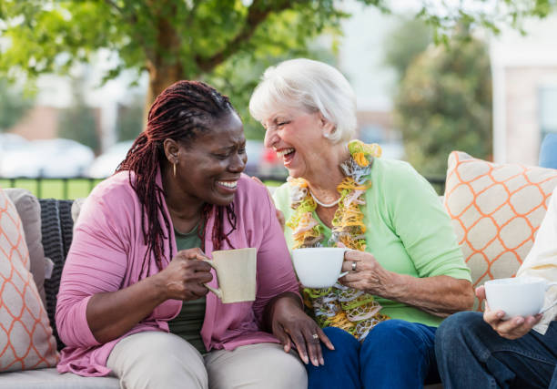 Senior woman, African-American friend laughing together A senior woman in her 70s, drinking coffee and laughing with her African-American friend, a mature woman in her 50s. They are sitting outdoors on a patio sofa. 55 59 years stock pictures, royalty-free photos & images