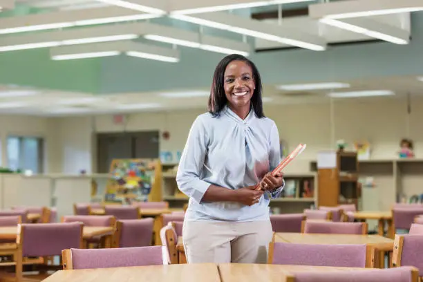 A mature African-American woman in her 40s standing in a library, smiling at the camera. She is a librarian or an elementary school teacher, carrying a book.