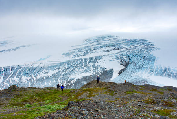 End of the trail. Tourists looking at the view. Closed clouds day, overlooking the Exit Glacier showing its blue ice, white snow, and a lush landscape. In Kenai Fjords National Park - July 27, 2018