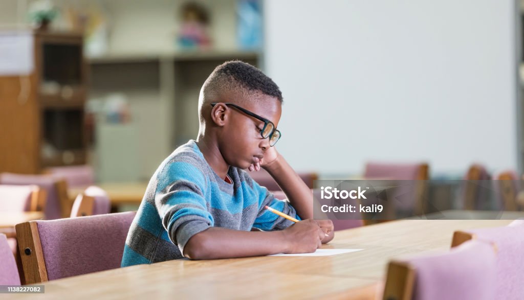 African-American boy in elementary school, writing A 10 year old African-American boy in elementary school, sitting alone at a table, writing with a serious expression on his face. He may be taking a test, or doing homework, or perhaps he was mischievous and is in school detention. Child Stock Photo