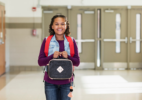 A 10 year old mixed race African-American and Caucasian girl attending elementary school. She is standing inside the school carrying a backpack and lunch box, smiling at the camera, happy to be back to school.