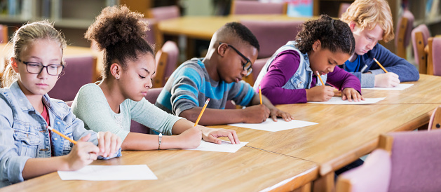 A group of five multi-ethnic elementary school students sitting in a row at a table, taking a test, writing with pencils on paper. They are 10 and 11 years old. The focus is on the African-American girl 2nd from the left.