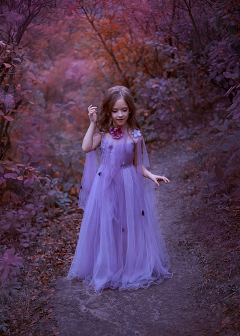 cute girl is standing in the forest in a purple light long dress with flowers, a little princess like in a dream, walks along the path in the maroon garden, straightens her brown hair, looks down.