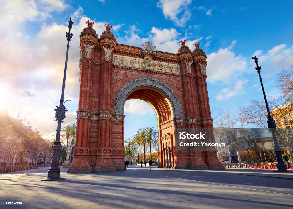 Sunrise at Triumphal Arch in Barcelona, Catalonia, Spain. Sunrise at Triumphal Arch in Barcelona, Catalonia, Spain. Arc de Triomf at boulevard street. Alley with tropical palm trees. Early morning landscape with shadows and blue sky with clouds. Famous Barcelona - Spain Stock Photo
