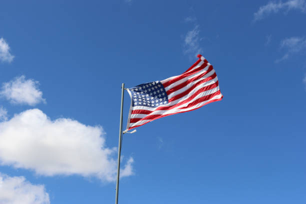 Stars and stripes national flag of the United States of America flying and fluttering on a grey pole with blue sky and white clouds in the background.