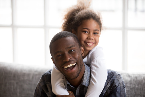 Headshot of smiling little African American girl piggyback young happy dad relaxing on couch together, portrait of excited black father embrace hug with small kid, posing for family picture at home