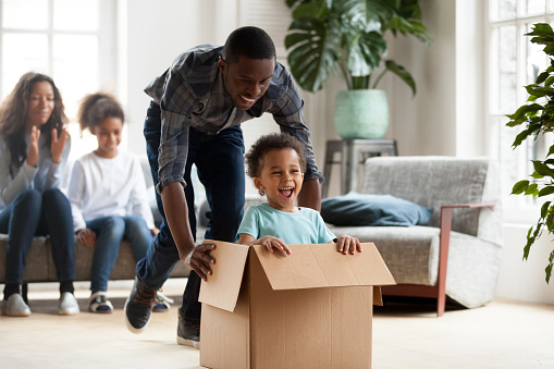 Excited little black boy sit in cardboard box playing with young dad unpacking packages on moving day, happy young African American family have fun entertain with small kids relocating to new home