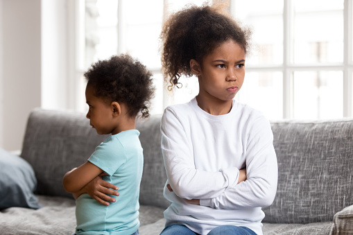 Stubborn small African American brother and sister sit on couch back to back avoid talking, offended mixed race siblings boy and girl with arms crossed angry after fight not speaking to each other