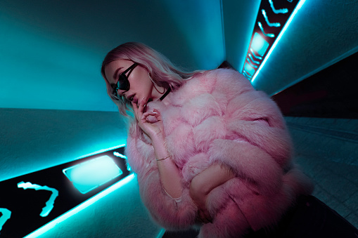 Teen hipster girl in stylish glasses and fur standing in blue neon lights elements on street wall, female teenager fashion model woman posing in city night turquoise