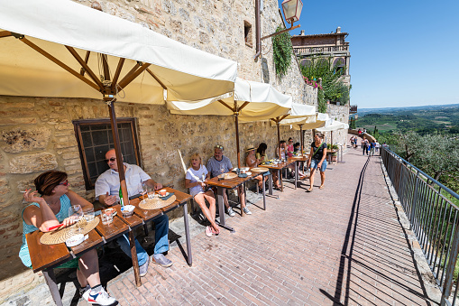 San Gimignano, Italy - August 27, 2018: People sitting drinking wine eating at cafe restaurant in historic town village in Tuscany during sunny summer day with view from hilltop