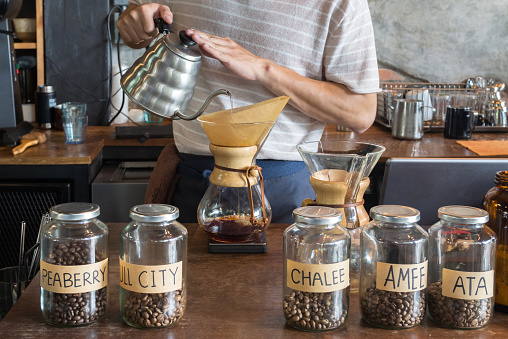 barista holding drip pot and pouring water onto ground coffee beans in the paper filter for brewing drip coffee at the cafe in the morning. coffee dripping is a pour-over method for filtering coffee.