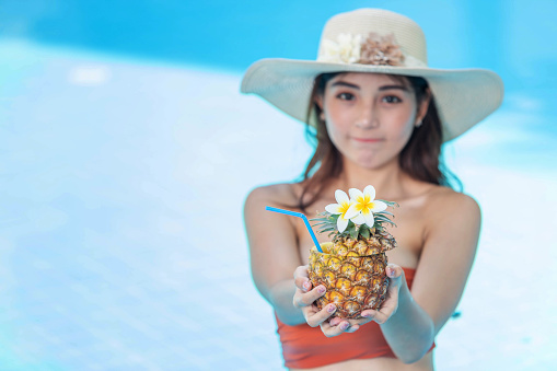 Beautiful female in bikini relaxing at the pool and holding pineapple cocktail.