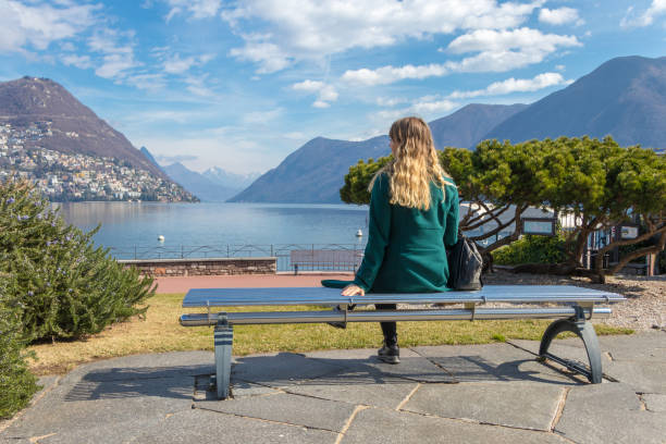 beautiful elegant woman sitting on a park bench and looking at lake lugano and alps mountains in switzerland - switzerland ticino canton lake lugano imagens e fotografias de stock