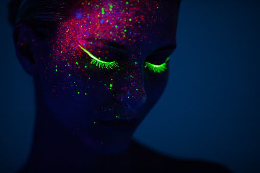 One young woman painted with fluorescent colors standing in front of ultraviolet light. Space is dark with blue background.
