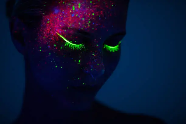 One young woman painted with fluorescent colors standing in front of ultraviolet light. Space is dark with blue background.