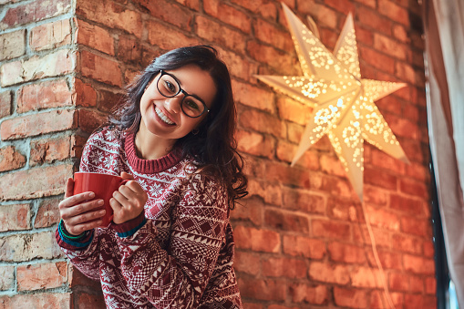 Christmas concept. Portrait of a cheerful brunette girl wearing eyeglasses and warm sweater holding a cup of coffee while leaning on a brick wall.
