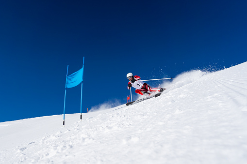 low angle view of male skier in race suit skiing giant slalom course on sunny winter day with clear blue sky snow splashing