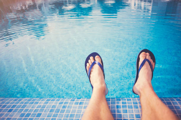 Man relaxing by blue swimming pool wearing sandals with stretched out legs - POV Point of view and selective focus