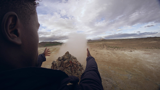 Young African ethnicity man dancing on a geothermal area. Catching warm steam. Geysers in background