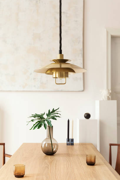 istock Eclectic and elegant dining room interior with design sharing table, chairs, gold pedant lamp, abstract paintings and stylish accessories. Tropical leafs in vase. Eclectic decor. Brown wooden parquet. 1138361152