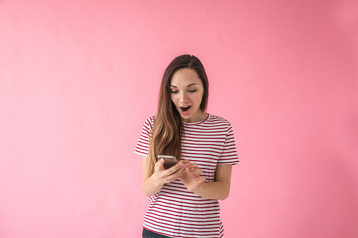 Surprised girl uses a cell phone on a pink background.