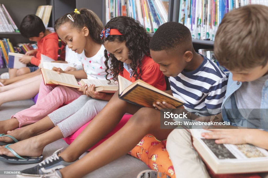 School kids sitting on cushions and studying over books in a library Side view of mixte ethnicity school kids sitting on cushions and studying over books in a library at school against bookshelfs in background Child Stock Photo