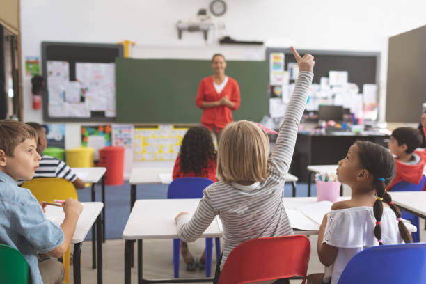Caucasian schoolgirl raising hand while his classmates next to her looking at her Rear view of a Caucasian schoolgirl raising hand while his classmates next to her looking at her with her teacher in background wavebreakmedia stock pictures, royalty-free photos & images