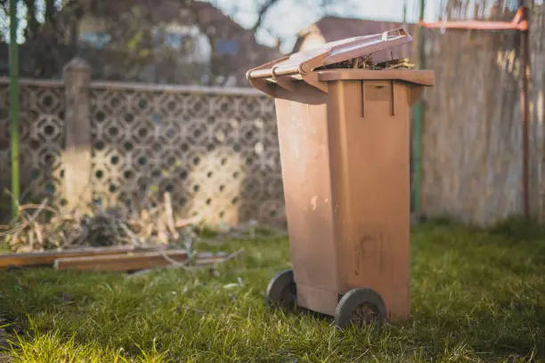 Brown trash bin for biodegradable waste on a garden as part of spring cleaning of a garden. Different parts of branches, leaves and grass are seen in the background.