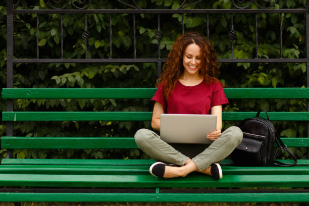 Portrait of a happy young woman smiling using laptop,sitting on bench in park. Lifestyle concept. Shooping online. stock photo