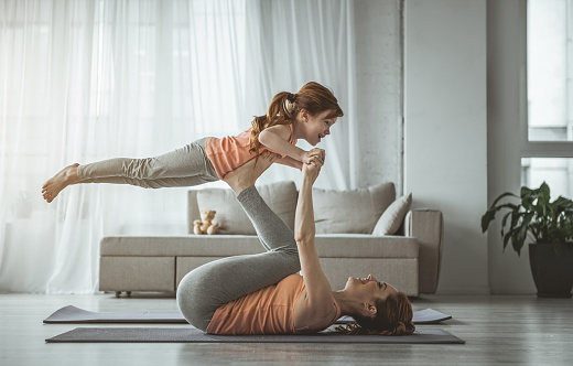 Little bird. Mother is lying on her back and holding her daughter on her feet during fitness at home. Girl looking happy while woman is laughing