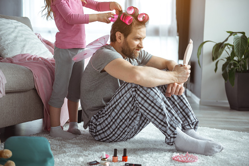 Cheerful daughter is sticking curlers on her father hair. Man is looking at mirror with shock while sitting on floor at home